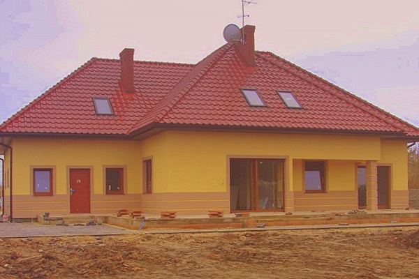 Żółty dom jednorodzinny z czerwoną dachówką, widoczne okna dachowe, komin i antena satelitarna, elewacja z poziomym pasem w innym odcieniu, teren wokół domu w trakcie prac wykończeniowych.